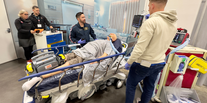 A medical transport crew in flight suits running a clinical simulation around a patient manikin with monitors and training carts