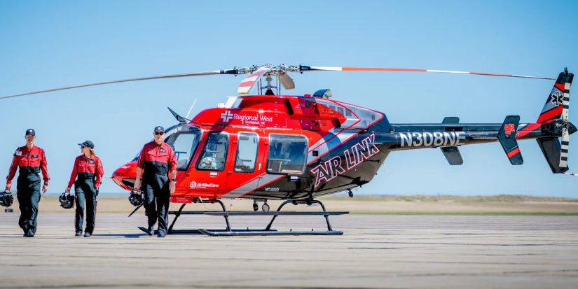 A flight crew in red and black flight suits carrying their helmets as they walk across a tarmac away from a medical helicopter